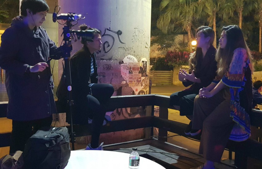 Two women sitting on high stools being interviewed by a film crew