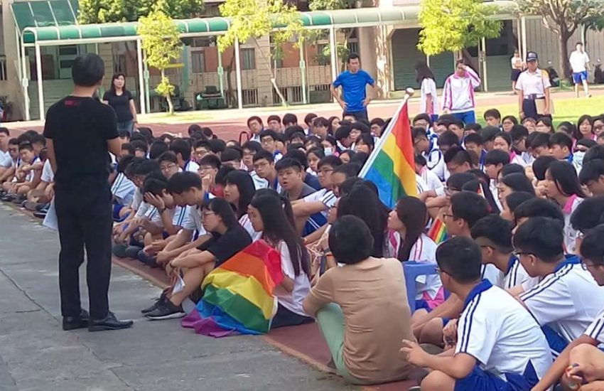 children sitting on the ground, some are draped in rainbow flags while others wave the flag on a flag pole