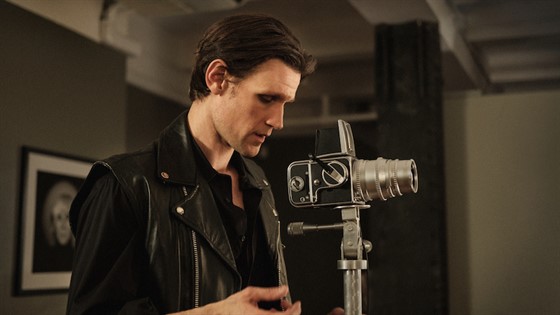 Actor Matt smith dressed in a black leather jacket looking into an old fashioned camera taking a photo
