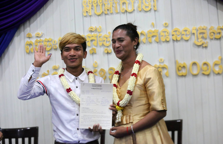 a couple stands holding a certificate and smiling. one of them is waving and they have a necklace of flowers around their necks