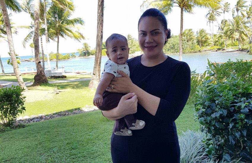 Hon. Frederica Tuita Filipe holds an infant on her hip she is wearing a black dress and they're standing outside in a tropical area