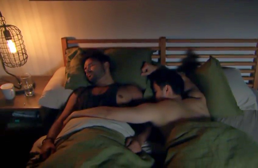 Two men lying in bed sleeping next to each other, one man has his arm across the other man's stomach