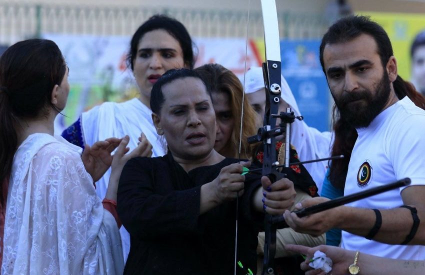 A trans woman in black preparing a bow and arrow to play archery being instructed by a man in a white t=shirt