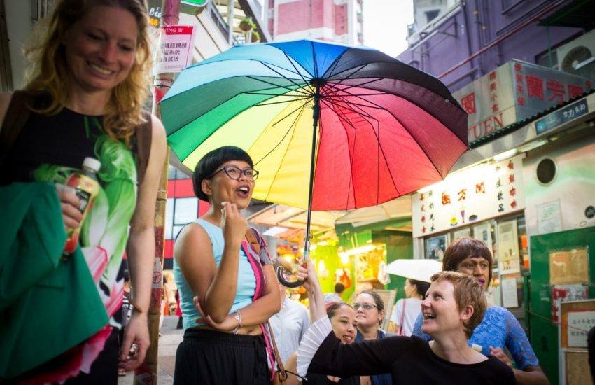 Three people standing on a step laughing, one is holding a rainbow umbrella over the heads of the others