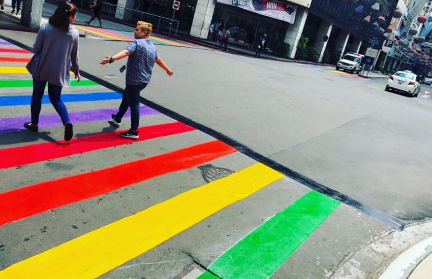 Two people walking across a rainbow crossing, the photos shows the backs of them 