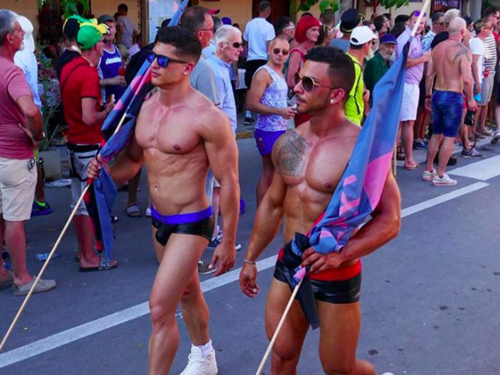 Two muscular men parade with flags during Sitges Pride