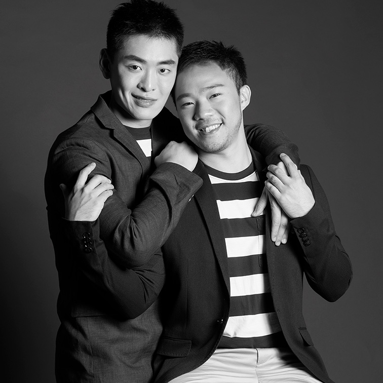 A black and white photo of a couple. One is standing behind the other who is sitting on a stool their arms are touching