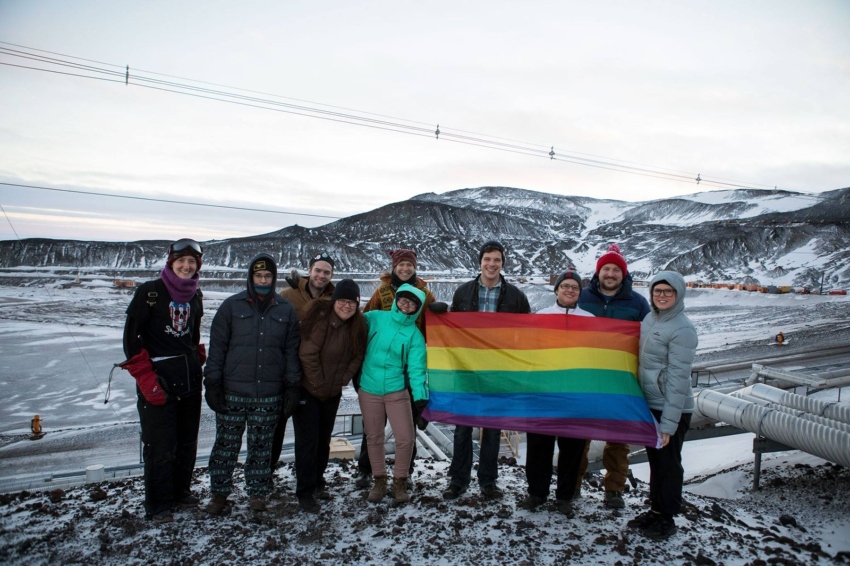 a group of people dressed up in winter clothes stand in front of a snowy location holding up a big pride rainbow flag