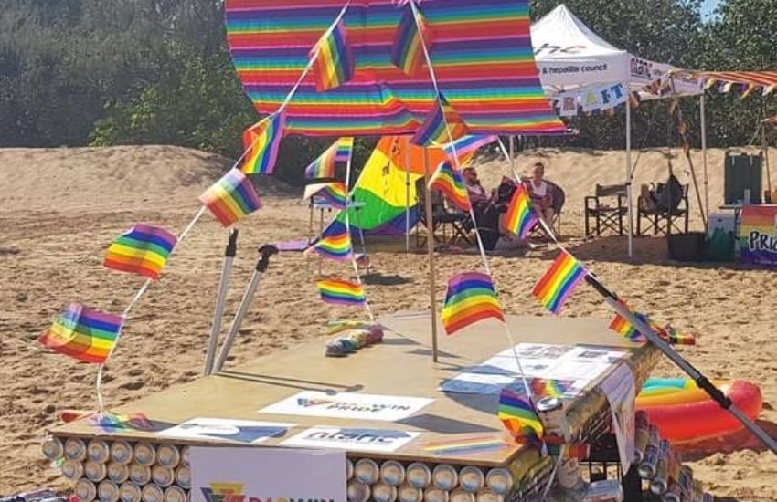 a raft made from beer cans and decorated in rainbow colours it is positioned on a sandy beach