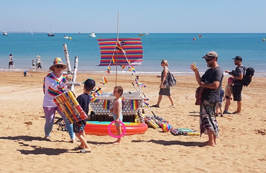 a wide shot facing the beach, on the sand is the rainbow raft and there are people standing around it it is a sunny day