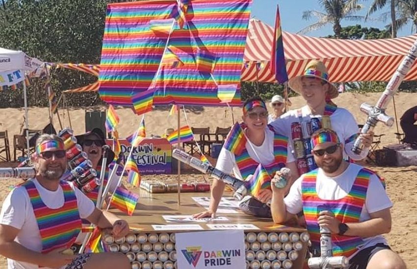 a group of people pose smiling next to a raft made from beer cans and decorated in rainbow colours