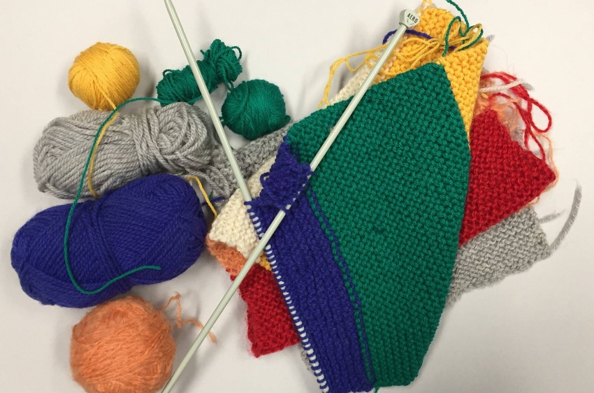 an overhead shot of knitting needles, wool and a colourful scarf against a white background