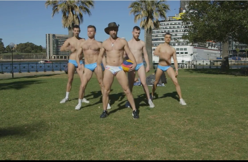 Five men wearing speedos stand in V formation, the man at the front is wearing an akubra hat and holding a water polo ball. they are outside at sydney's circular quay on a sunny day