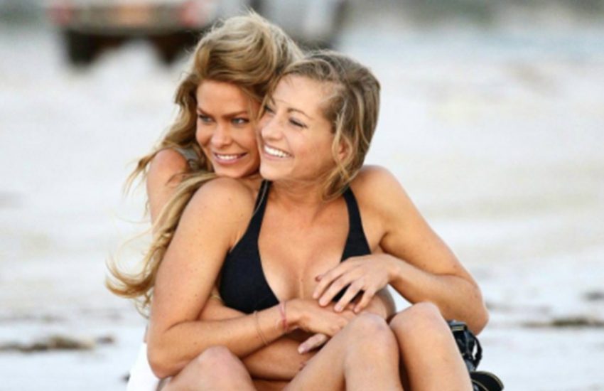 Megan Marx and Tiffany James in happier times in bikinis on a beach with sitting wrapped around each other