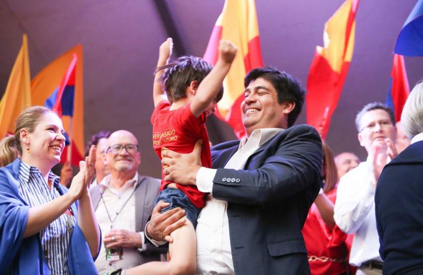 Carlos Alvarado on stage smiling holding a child throwing his arms in the area surround by flags and people cheering