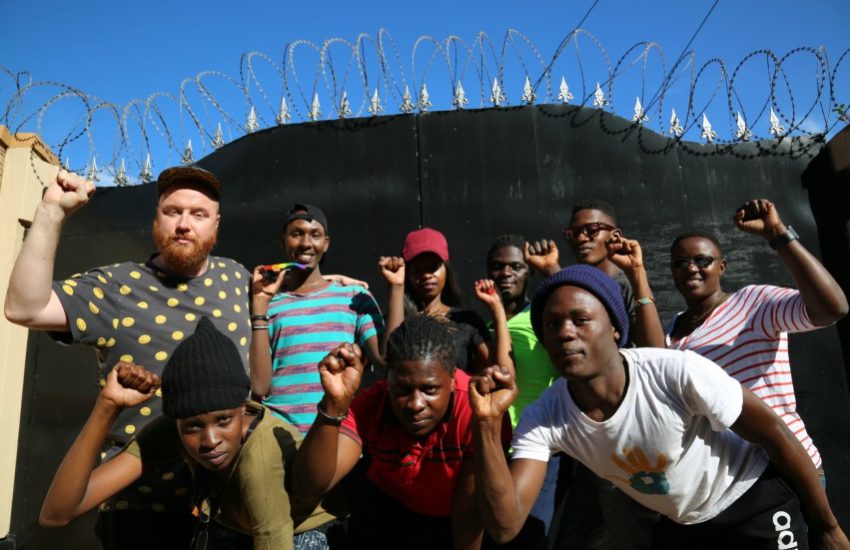 A group of people leaning into a camera raising their rights hands into a fist. it is a sunny day and there is barbed wire on a fence behind them