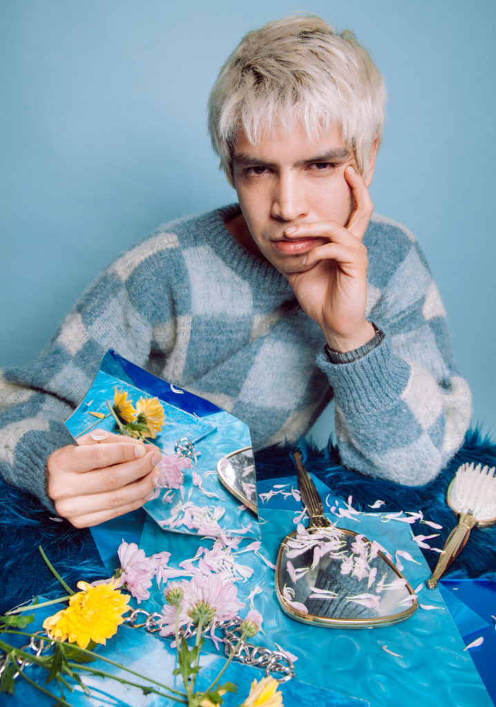 julio torres sits at a small table covered with a blue tablecloth holding broken flowers, his chin rests in one of his hands and he looks at the camera sultrly