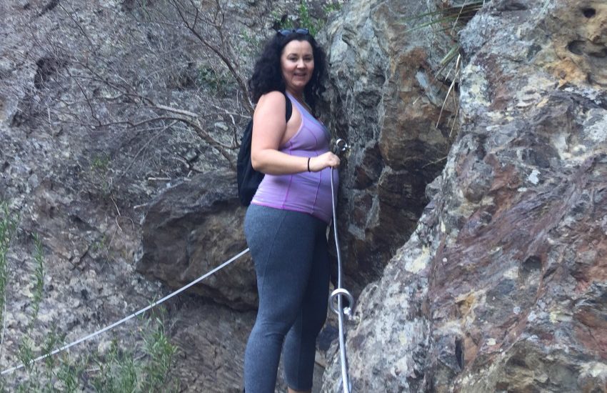 Photo taken upwards of shannon who is scaling a rock face. she is holding a steel cable in a purple gym top and smiling down over her right shoulder at the camera