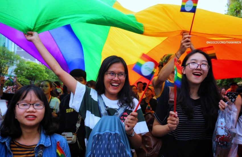 people standing under a horizontal rainbow flag they are smiling as they hold up the flag