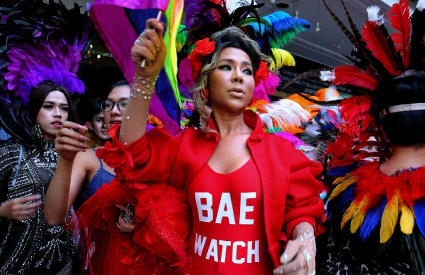 a drag queen in a red swimsuit that reads 'bae watch' she is waving a small rainbow flag behind her head