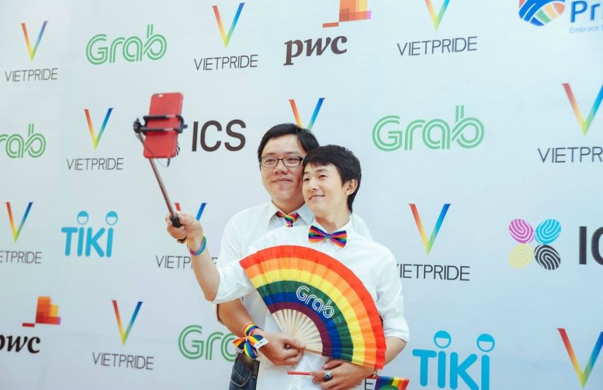 a couple taking a selfie with a selfie stick on the red carpet one is holding open a rainbow flag