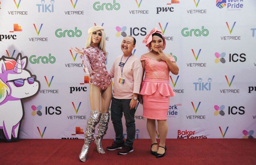 two women (possibly drag queens) in pink outfits on the red carpet with a man in a pink v-knit jumper